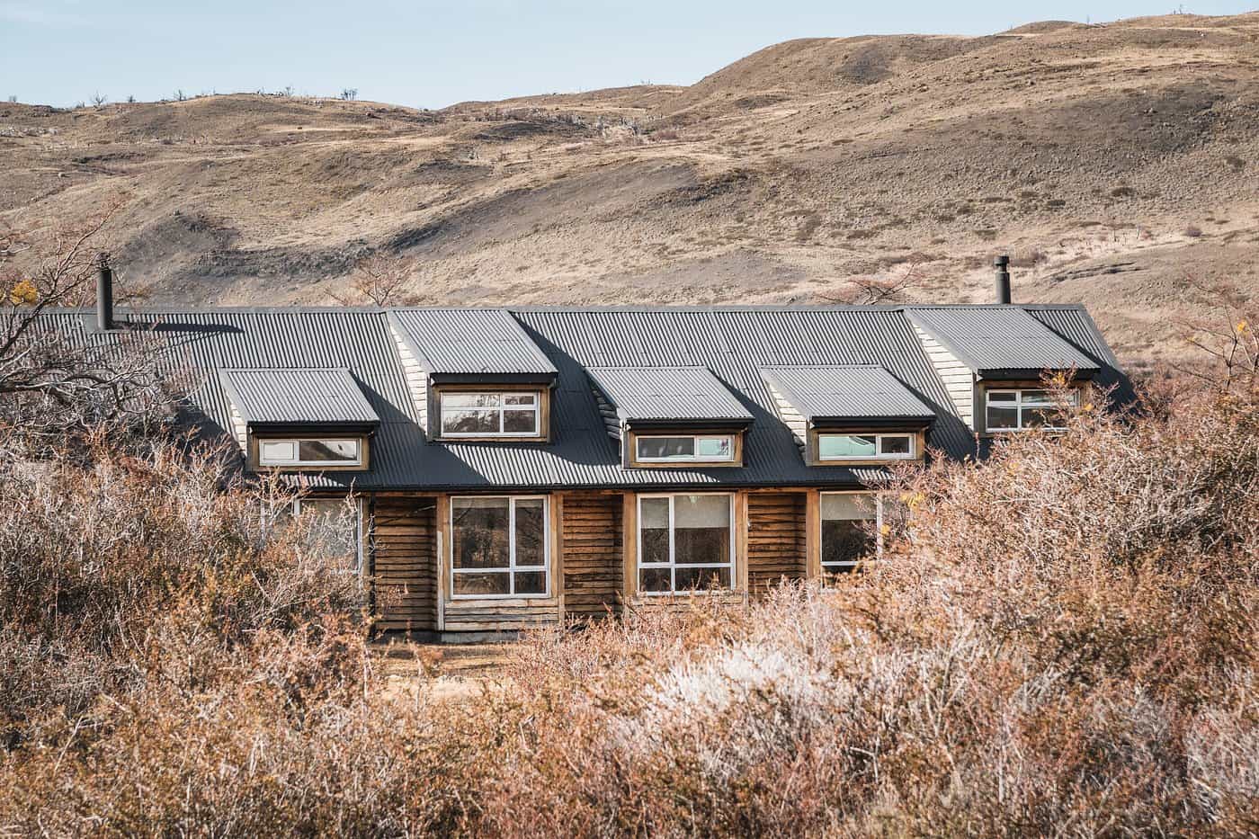 Refugio Centra Los Torres log cabin partially hidden by dry brush against arid hills.