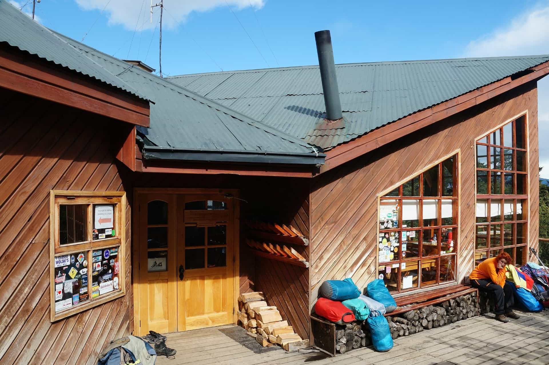 Wooden refuge or cabin exterior with outdoor gear, firewood, and a person resting outdoors.