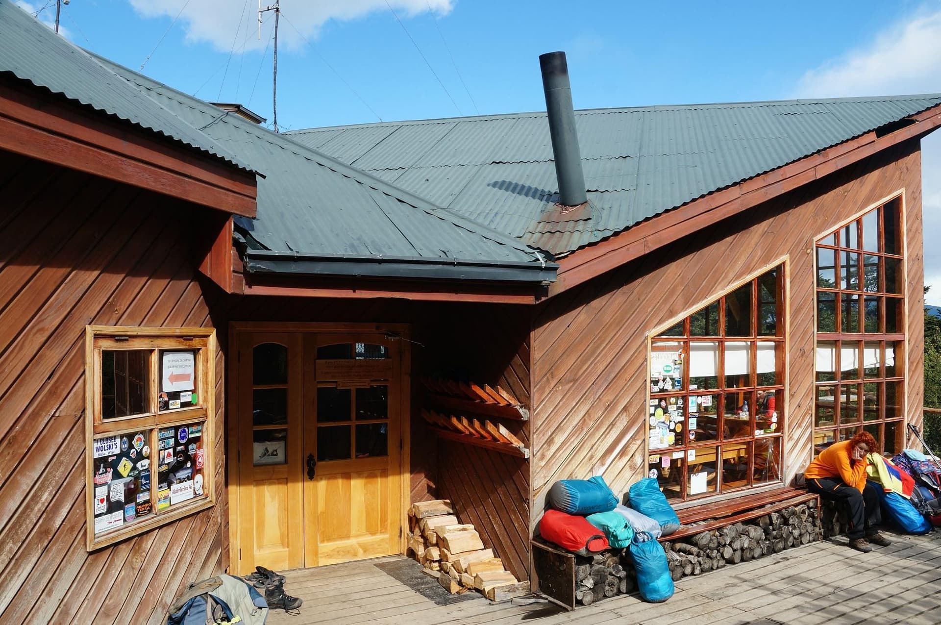 Wooden refuge or cabin exterior with outdoor gear, firewood, and a person resting outdoors.