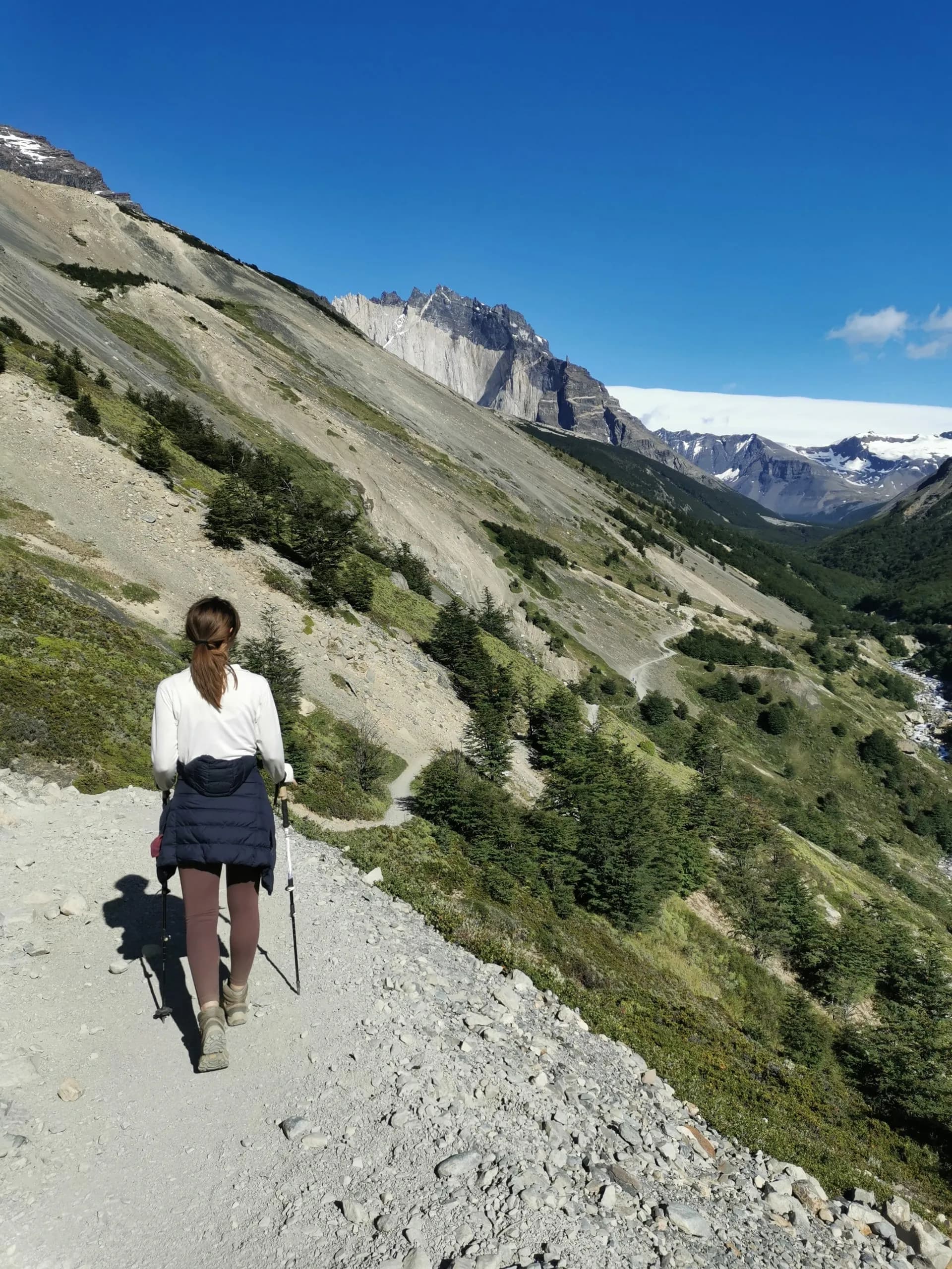 Hiker with trekking poles walking on rocky trail toward jagged, snow-capped mountains under blue sky.