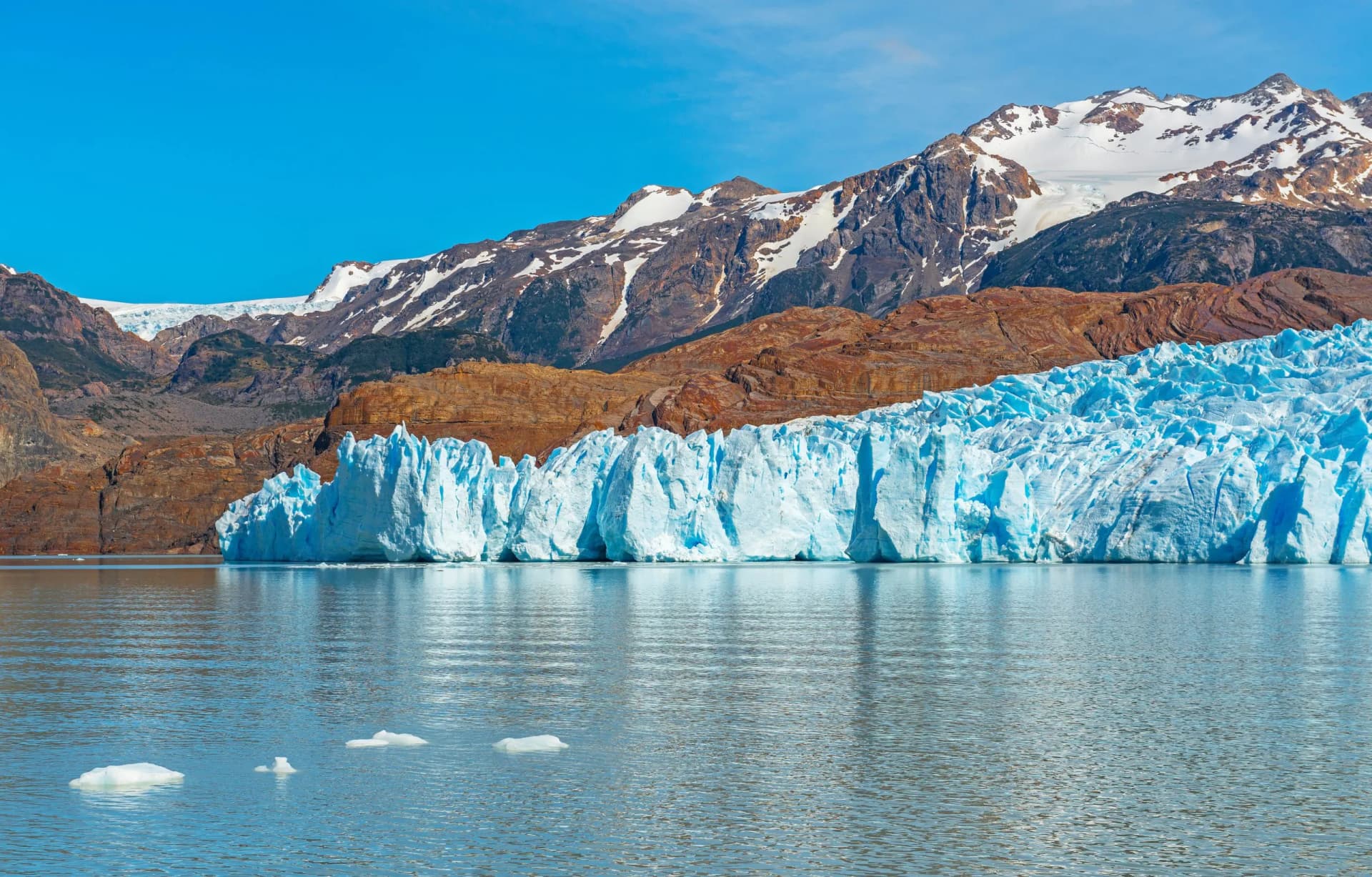 Glacier calving into water with snow-capped mountains and blue sky in the background