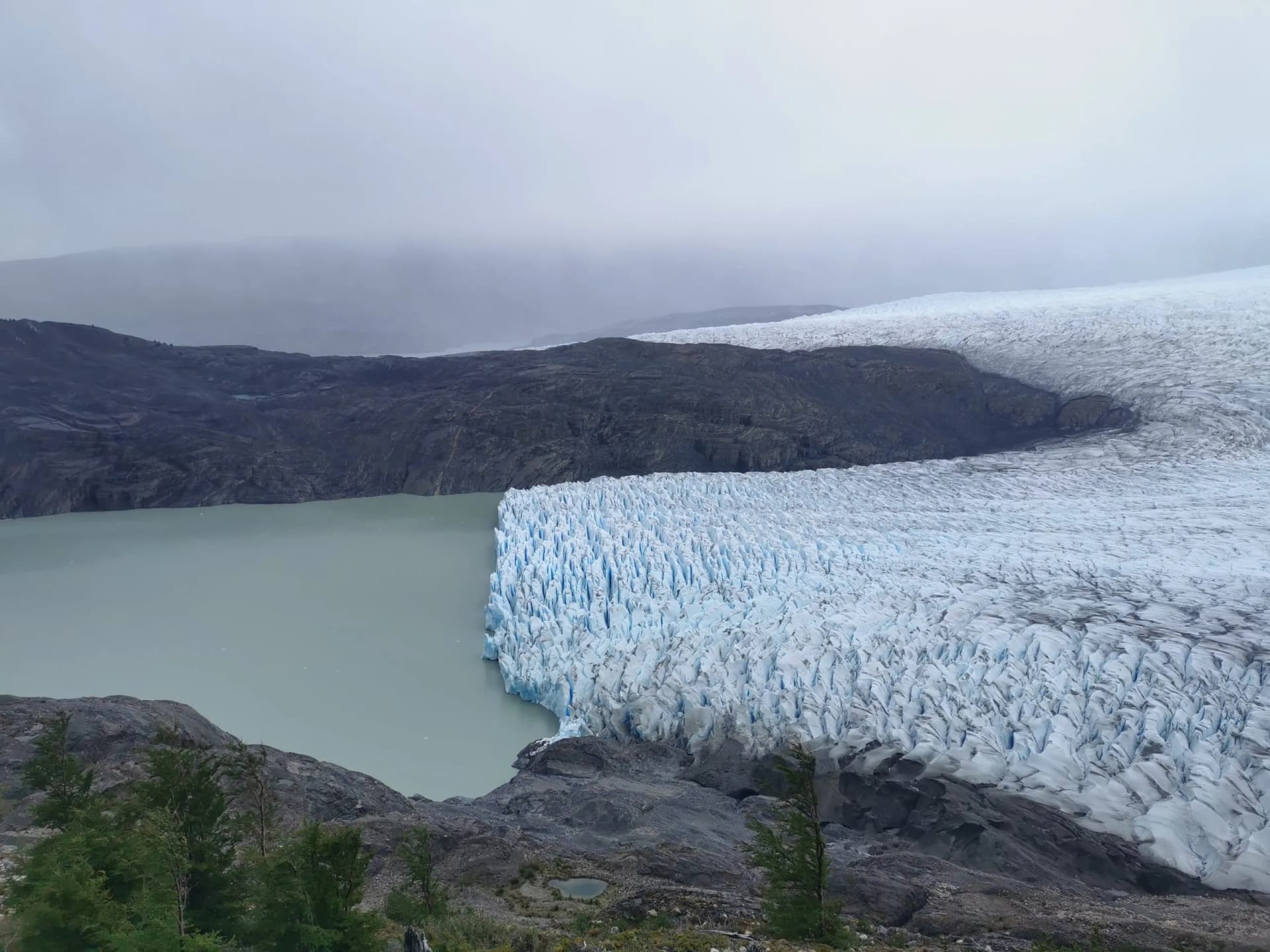 Glacier terminus meeting milky lake water, surrounded by dark mountains under a cloudy sky.