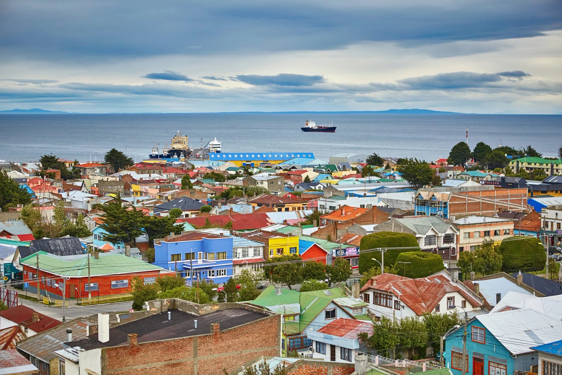 Colorful coastal town overlooking the Magellan Strait with ships docked and sailing under a cloudy sky.