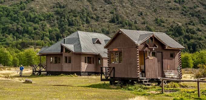 Two brown wooden cabins in a grassy clearing before a steep, forested hill.